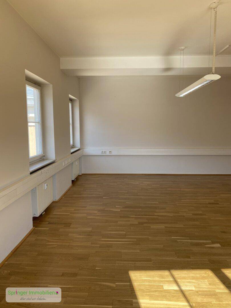 Leerer Büroraum mit Holzparkett, weißen Wänden und Heizkörpern unter den Fenstern.