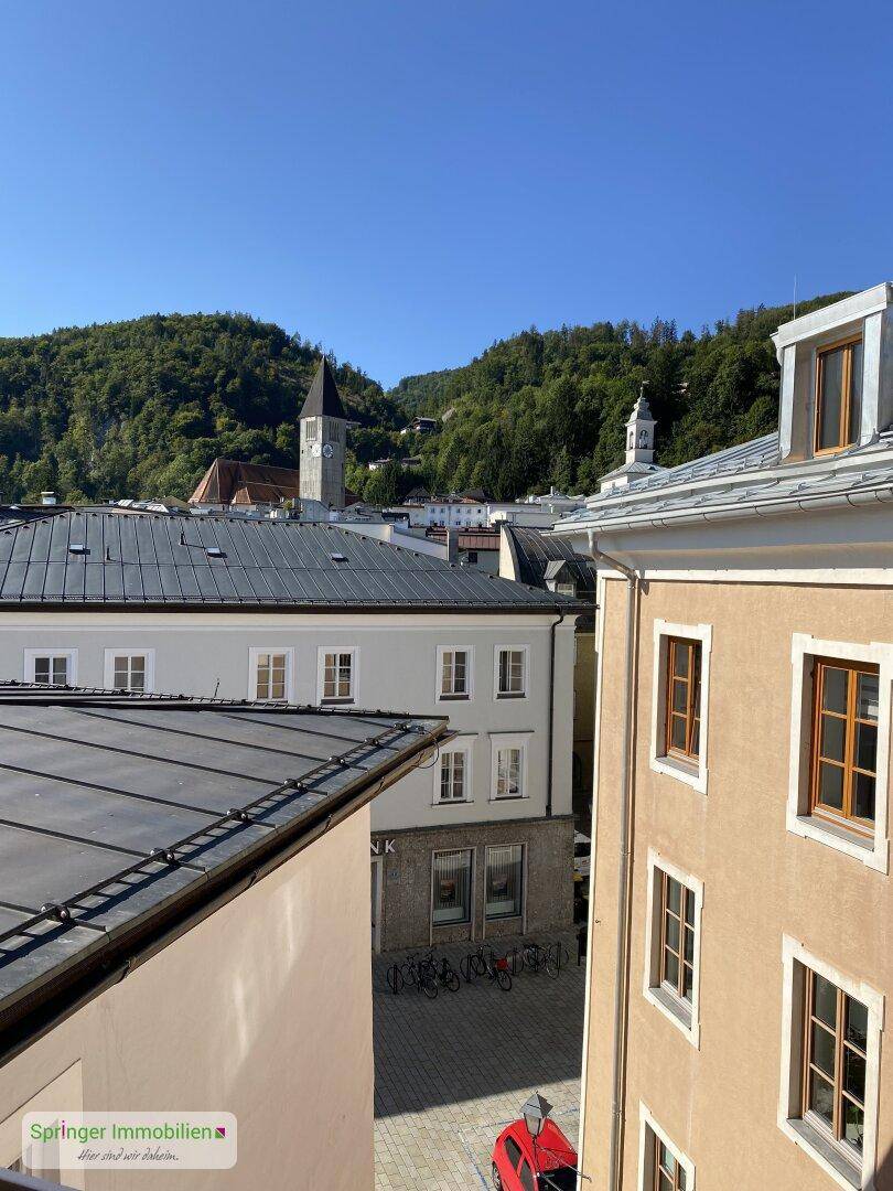 Panoramablick über die Dächer der Stadt mit grünen Hügeln und einem Kirchturm im Hintergrund.