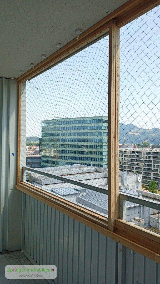 Blick aus einer verglasten Loggia mit Schutznetz auf die umliegenden Gebäude und die Stadt.