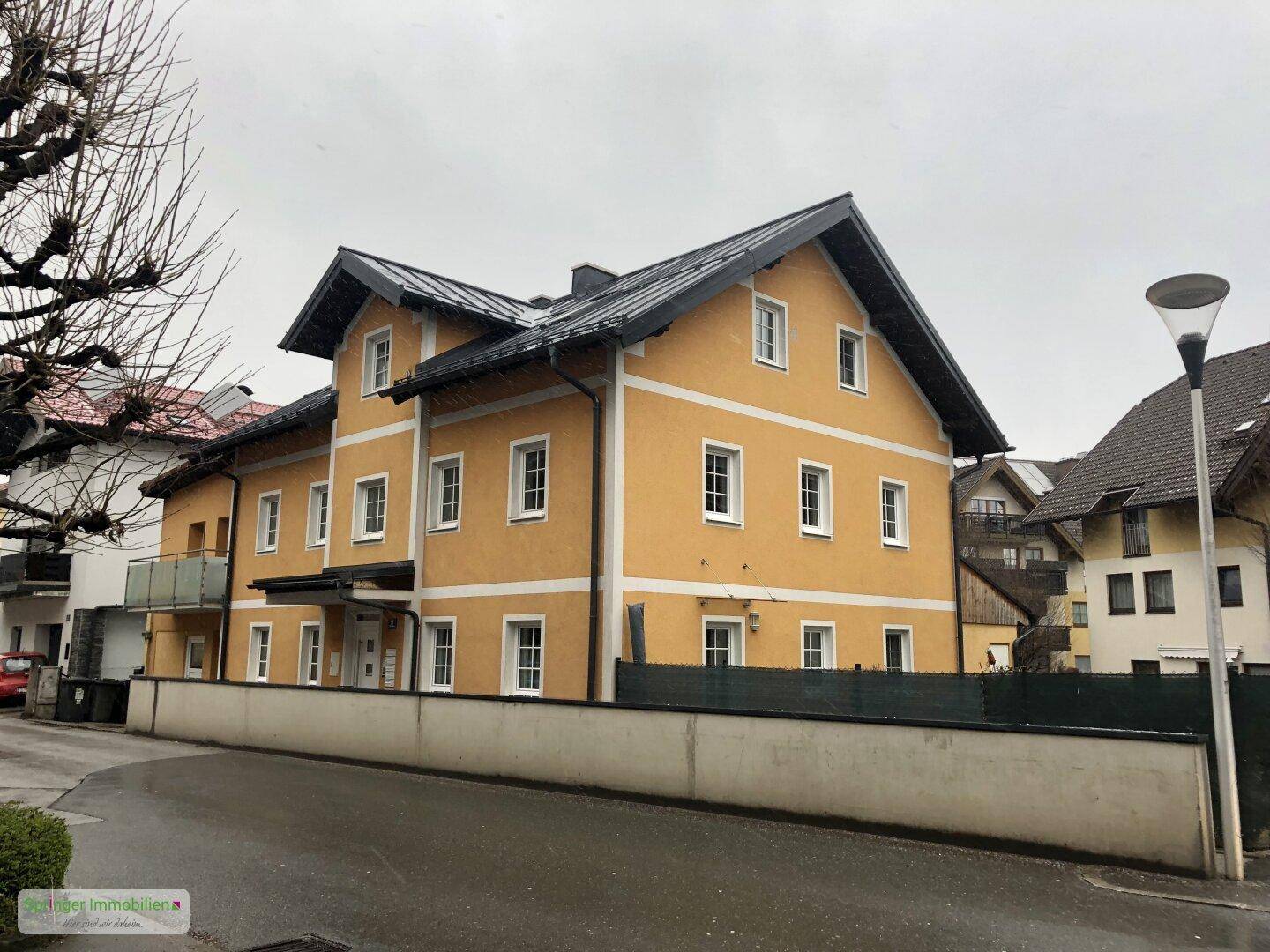 Gelbes Mehrfamilienhaus mit dunklem Dach und weißen Fenstern an einer ruhigen Straße gelegen.