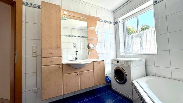 Gefliestes Badezimmer mit Waschtisch, Spiegelschrank, Badewanne und Waschmaschine.