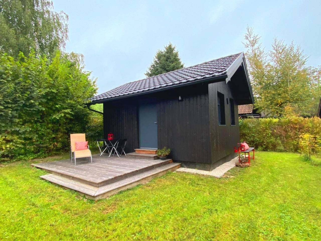 Kleines schwarzes Holzhaus mit Terrasse und Garten, umgeben von Bäumen und Sträuchern.