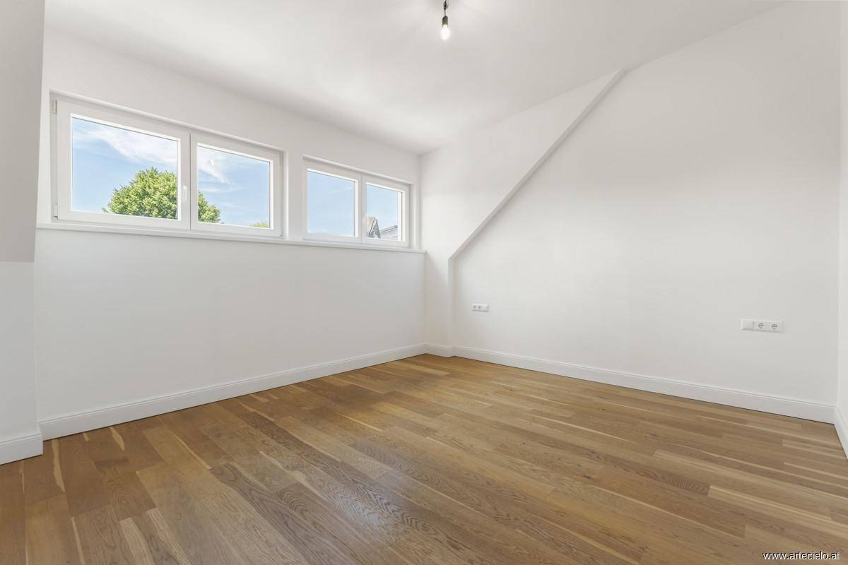 Helles Zimmer mit Holzboden, großen Fenstern und schrägen Wänden im Dachgeschoss.