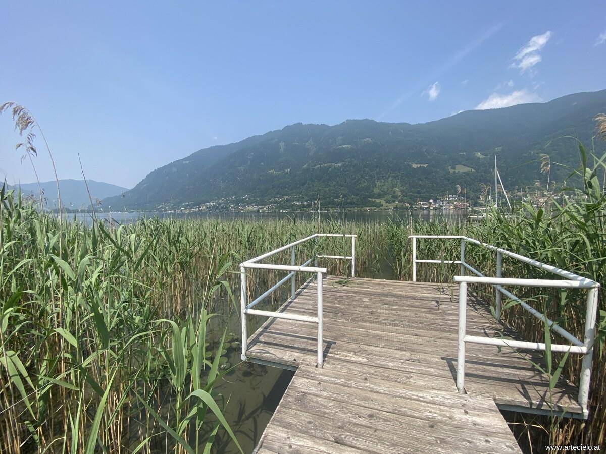 Holzsteg umgeben von Schilf mit weitem Blick auf den See und die Berge.