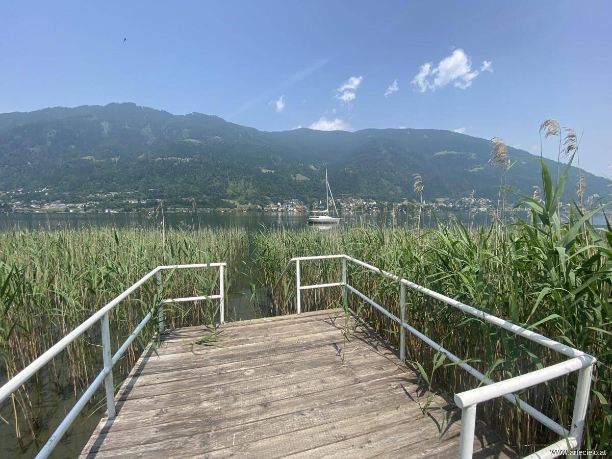 Breiter Blick vom Holzsteg über den See mit Schilf und majestätischen Bergen im Hintergrund.