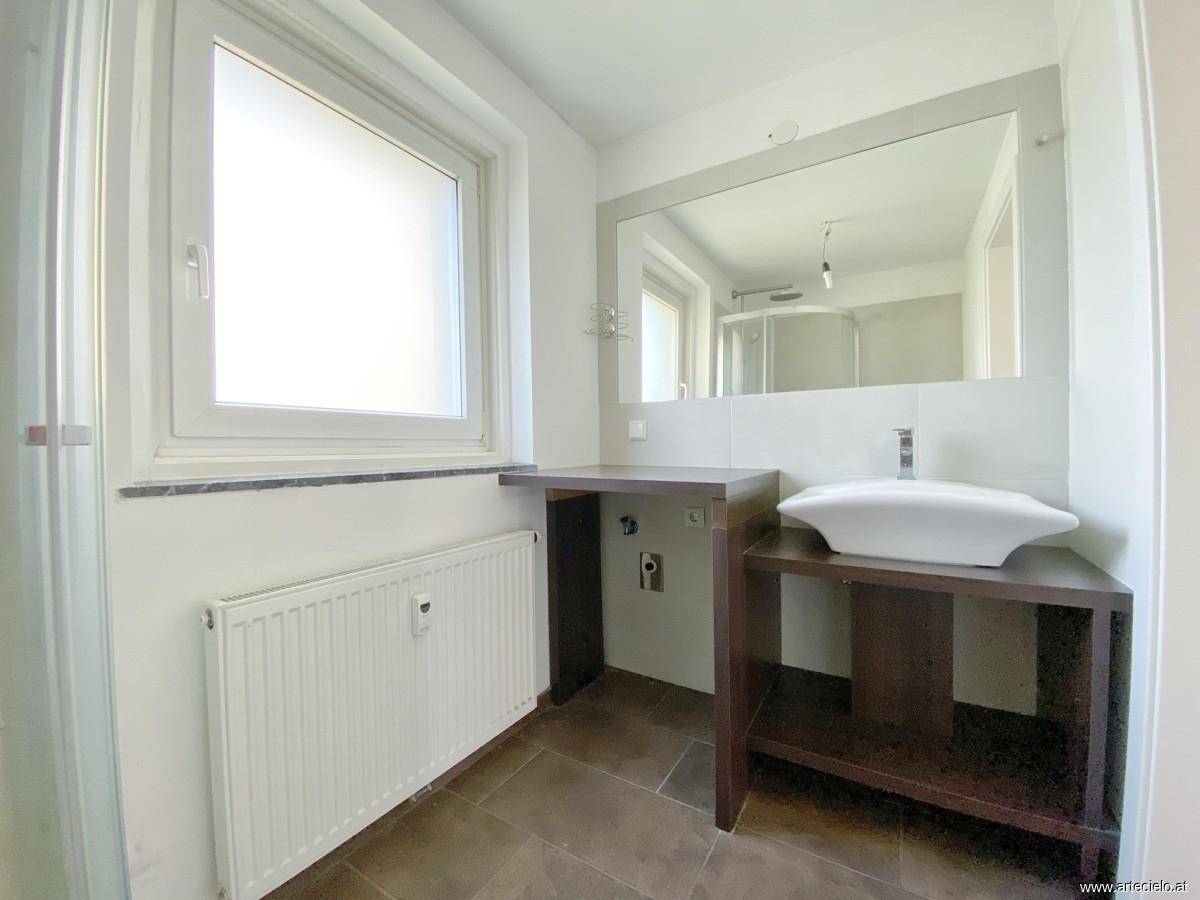 Modernes Badezimmer mit Waschtisch, großem Spiegel und Fenster, das viel Tageslicht hereinlässt.