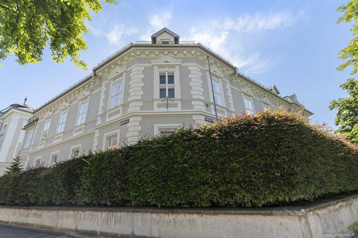 Elegantes Eckgebäude mit gepflegter Hecke und klassischer Fassadengestaltung unter blauem Himmel.