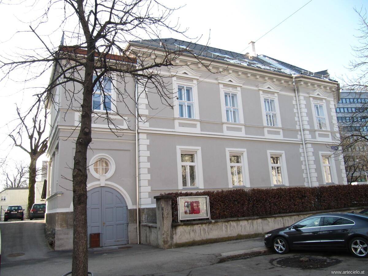 Historisches Gebäude mit grauer Fassade und weißen Akzenten, umgeben von kahlen Bäumen im Winter.