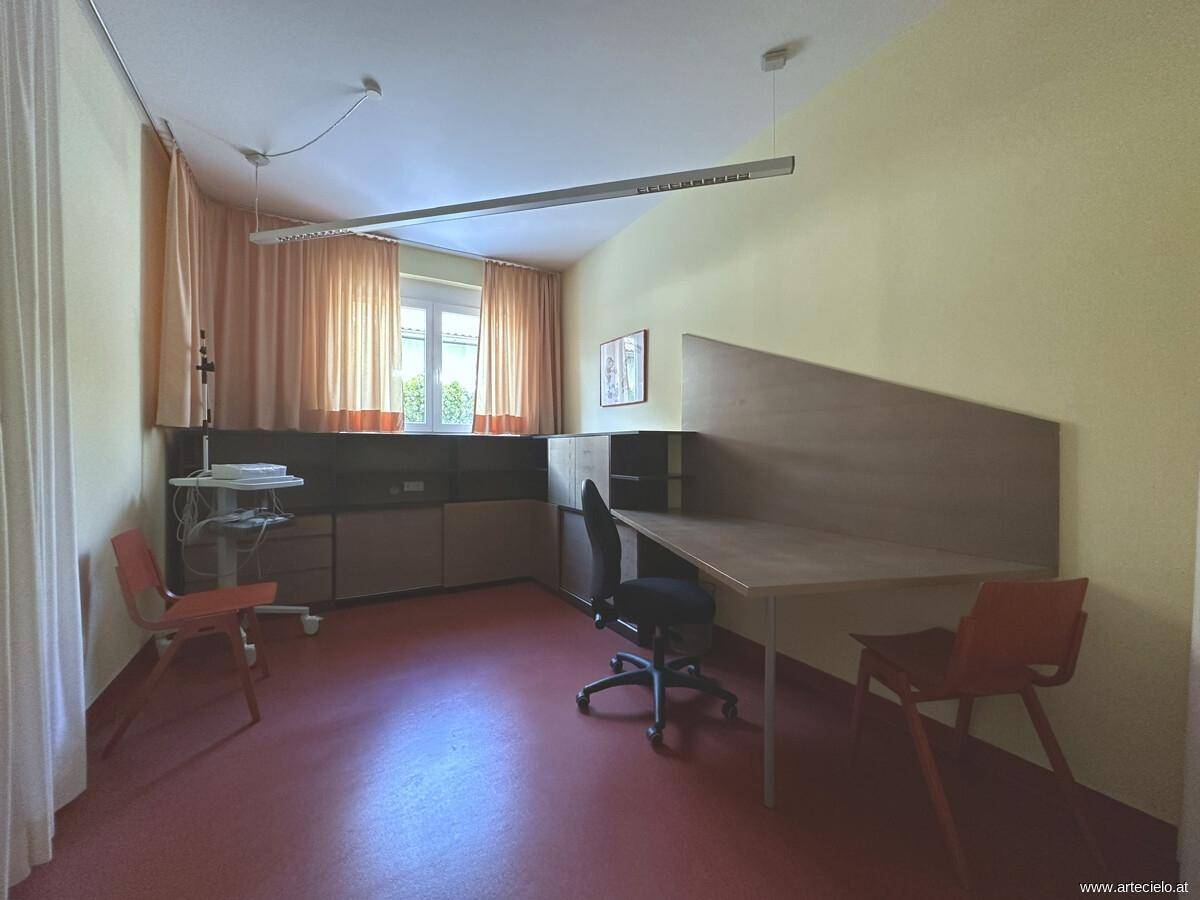 Funktionaler Büroraum mit Schreibtisch, Stühlen, medizinischer Ausrüstung und Vorhängen am Fenster.