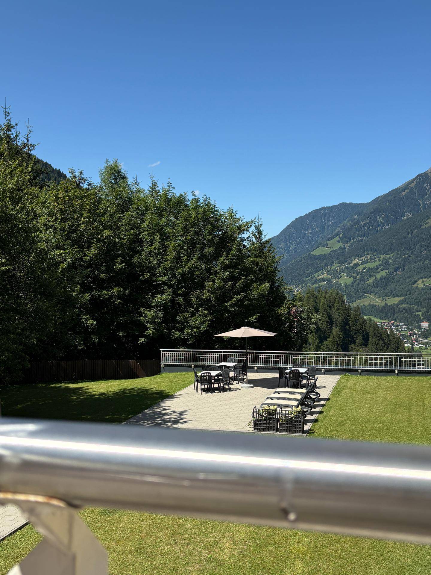 Weitläufige Gartenanlage mit Sonnenterrasse, Liegestühlen und beeindruckendem Bergblick unter blauem Himmel.