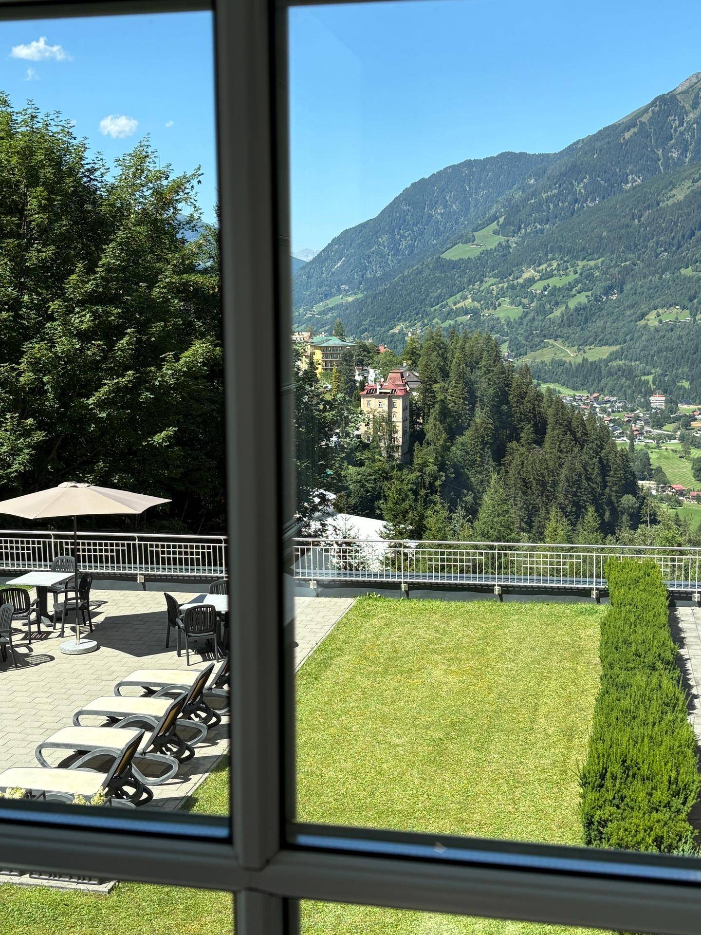 Blick aus einem Fenster auf eine gepflegte Rasenfläche, eine Terrasse mit Liegestühlen und die umliegende Berglandschaft.