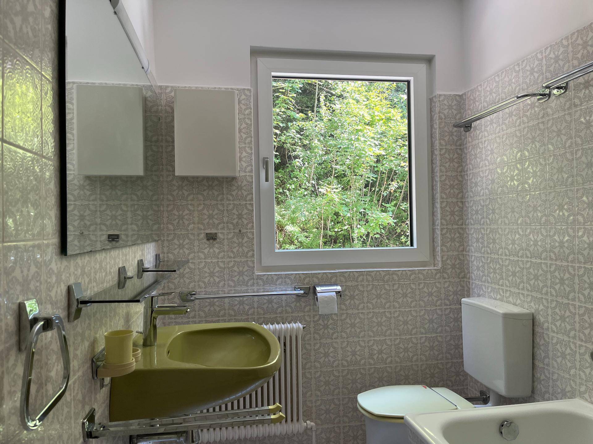 Badezimmer mit Fenster, grünem Waschbecken, Toilette und gemusterten Fliesen an den Wänden.