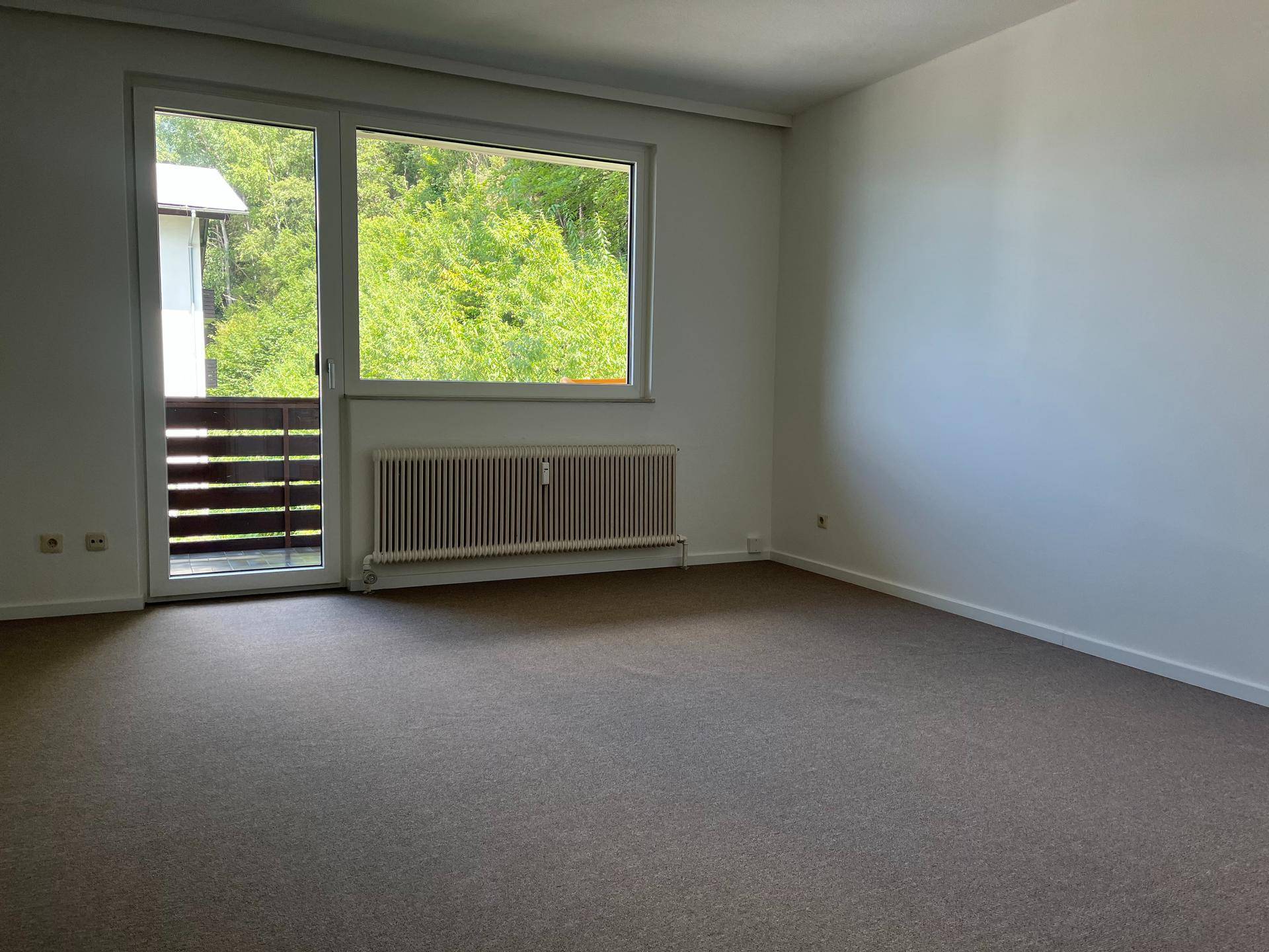 Heller, leerer Wohnraum mit Teppichboden, großem Fenster und Zugang zum Balkon.
