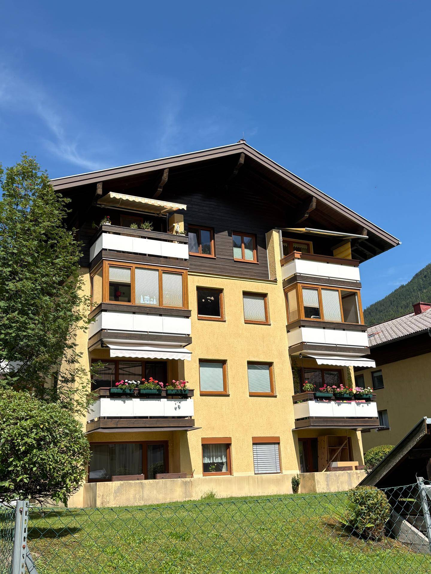 Mehrfamilienhaus mit gelber Fassade, Holzbalkonen und Satteldach in alpiner Umgebung.