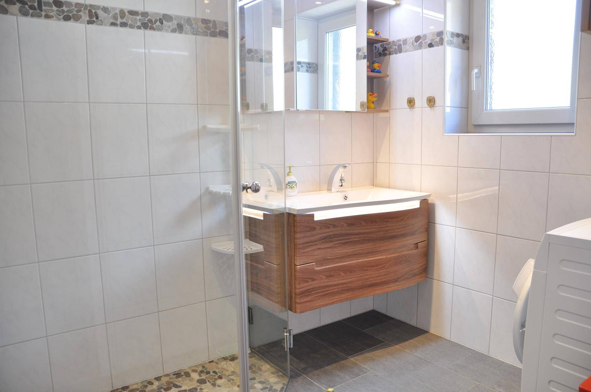 Modernes Badezimmer mit Duschkabine, schwebendem Holzwaschtisch und Fenster für Tageslicht.