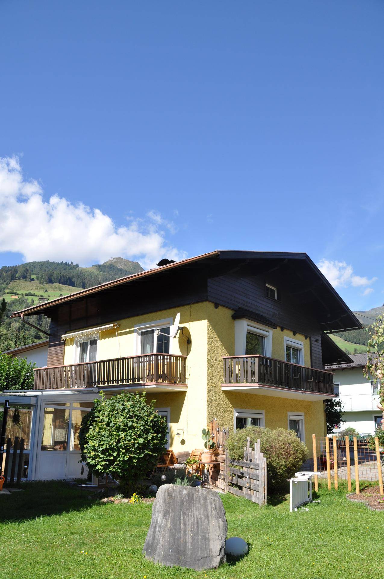Gelbes Einfamilienhaus mit mehreren Balkonen, gepflegtem Garten und Bergblick im Hintergrund.