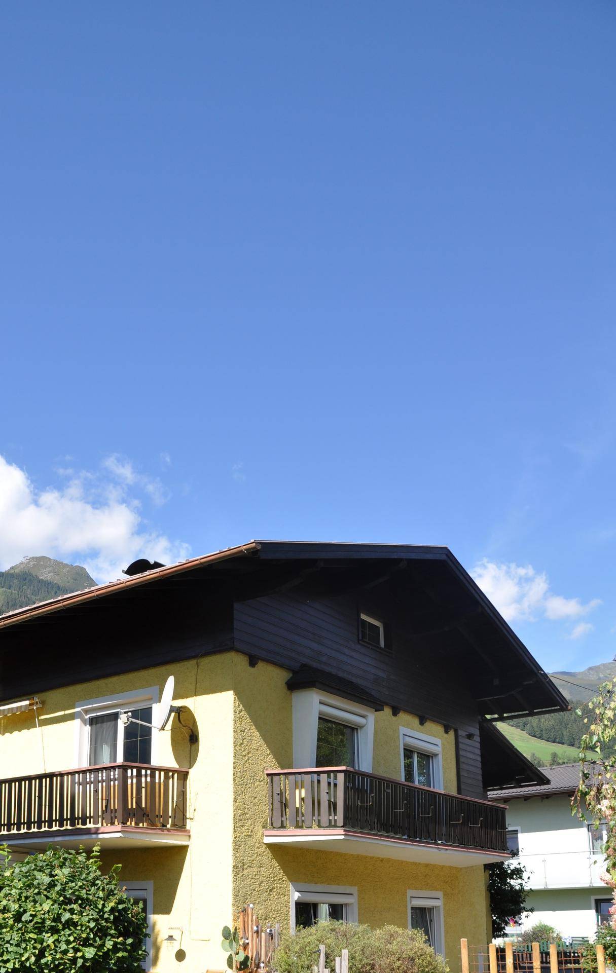 Gelbes Haus mit traditionellen Balkonen und Blick auf die umliegenden Berge unter blauem Himmel.