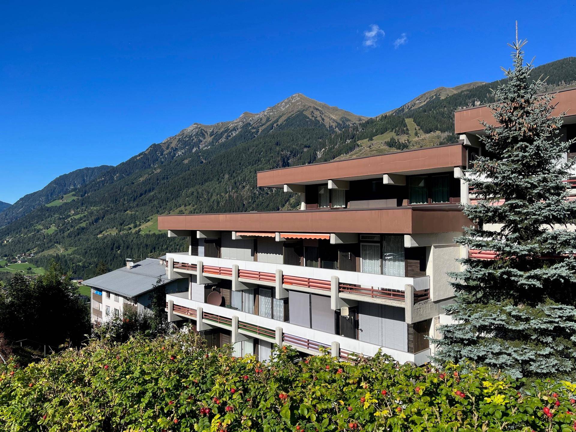 Mehrfamilienhaus in alpiner Umgebung mit Balkonen und beeindruckendem Bergblick unter blauem Himmel.