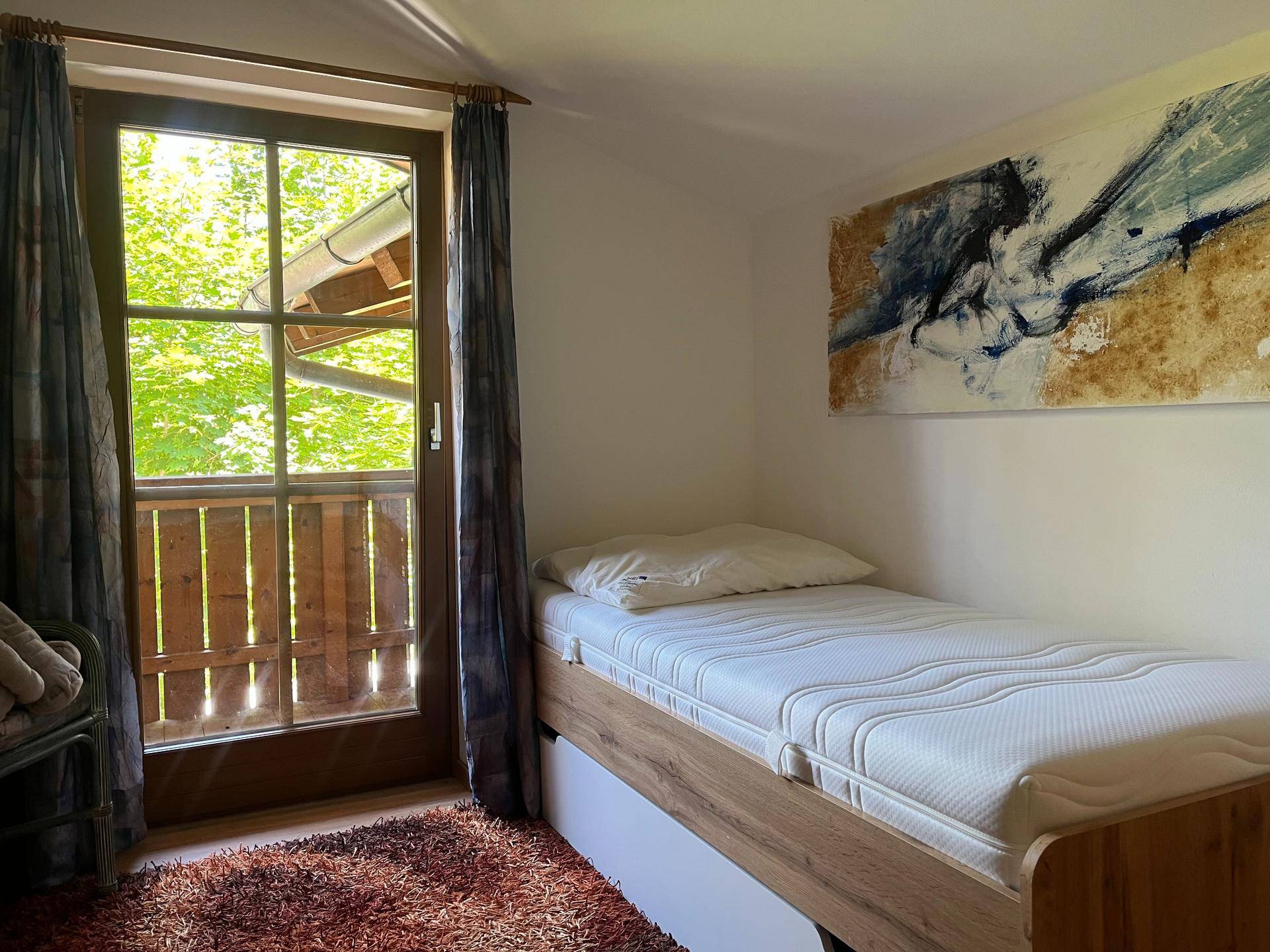 Helles Schlafzimmer mit Einzelbett, Holzbalkon und einem schönen Ausblick ins Grüne.