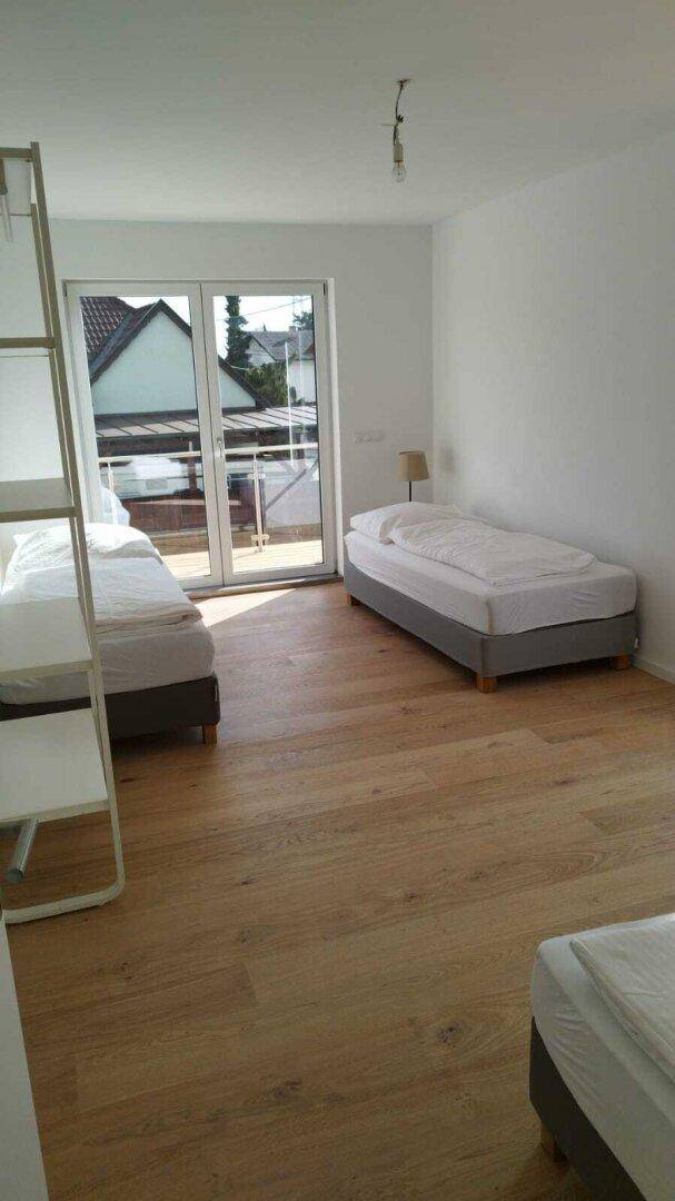 Helles Schlafzimmer mit Holzboden, mehreren Betten und Zugang zu einem kleinen Balkon.