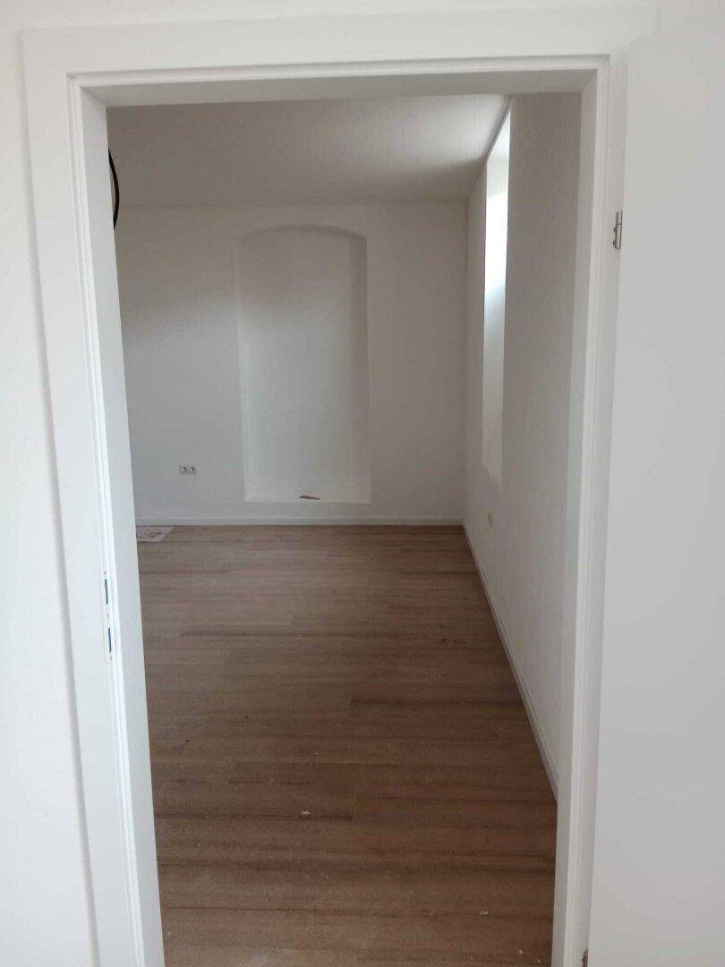 Heller, leerer Raum mit hellem Holzboden und weißen Wänden, gesehen durch eine Türöffnung.