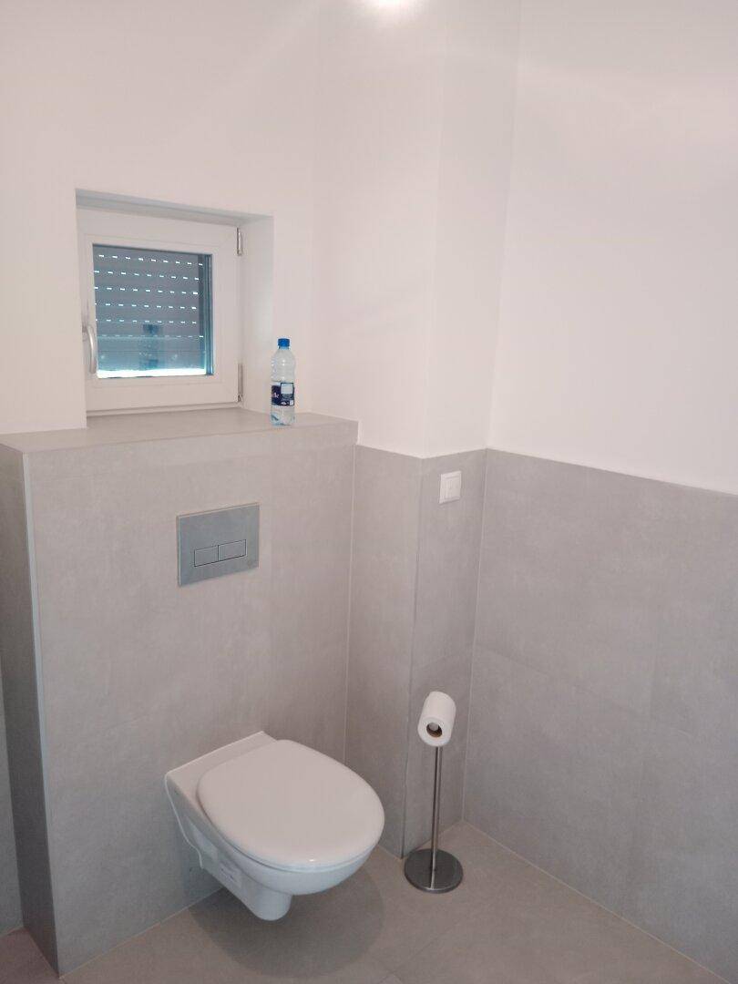 Modernes Gäste-WC mit wandhängender Toilette, grauen Fliesen und einem kleinen Fenster mit Rollladen.