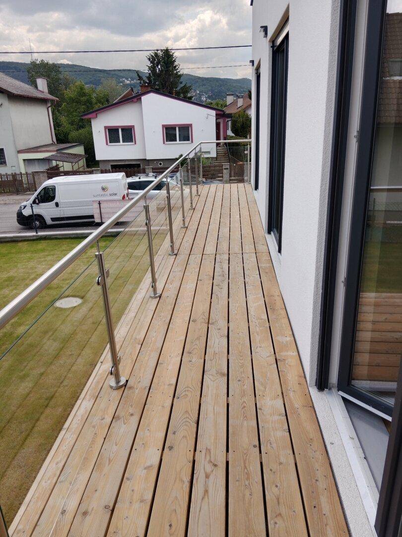 Geräumiger Holzbalkon mit Glasgeländer und Blick auf den Garten und die Umgebung.