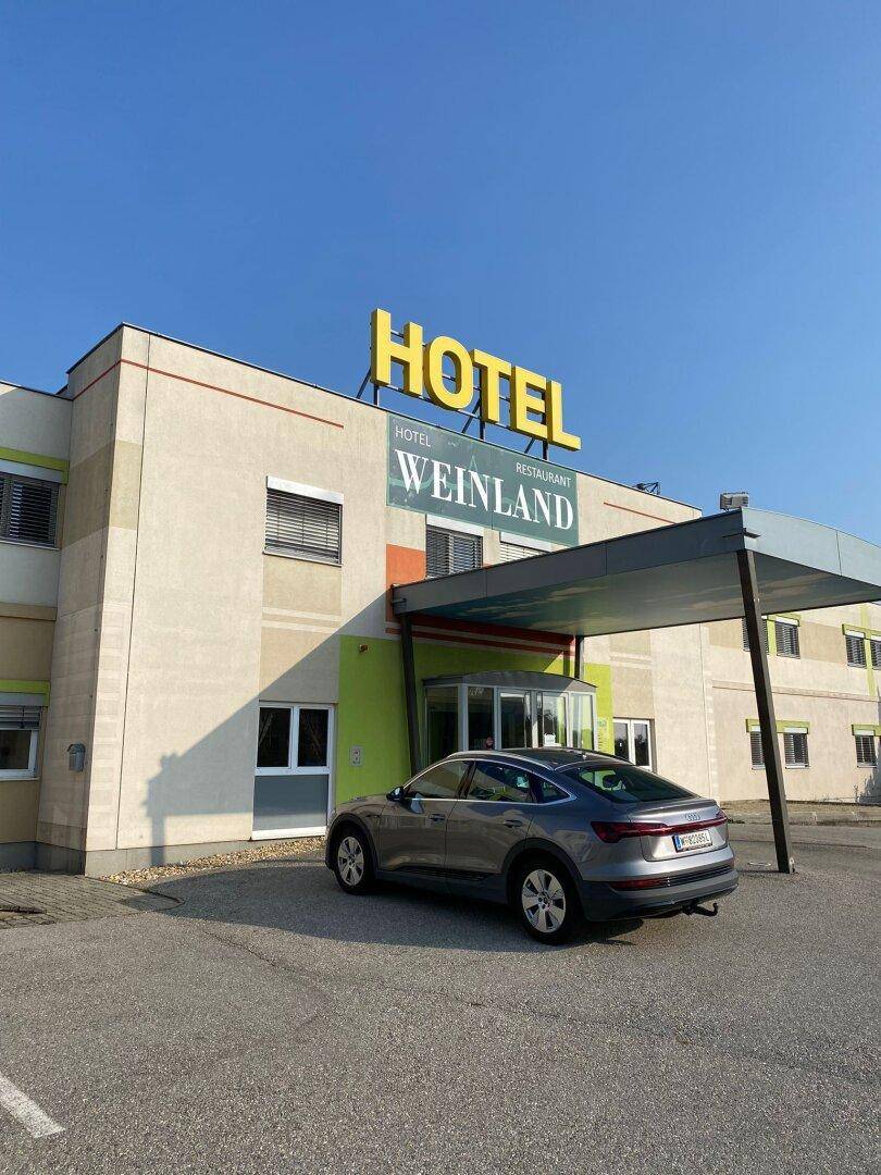 Außenansicht eines modernen Hotels mit dem Namen "Hotel Weinland" und einem geparkten Elektrofahrzeug davor.