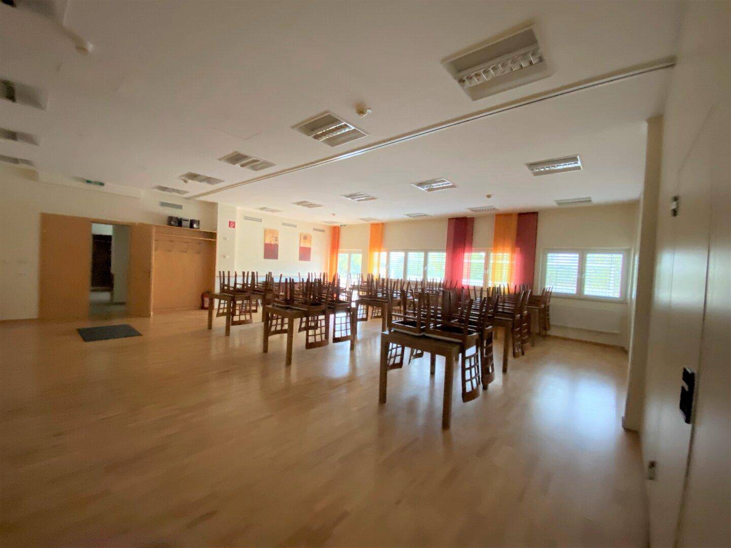 Geräumiger Saal mit Holzboden und vielen Tischen und Stühlen, ideal für größere Versammlungen oder Feierlichkeiten.