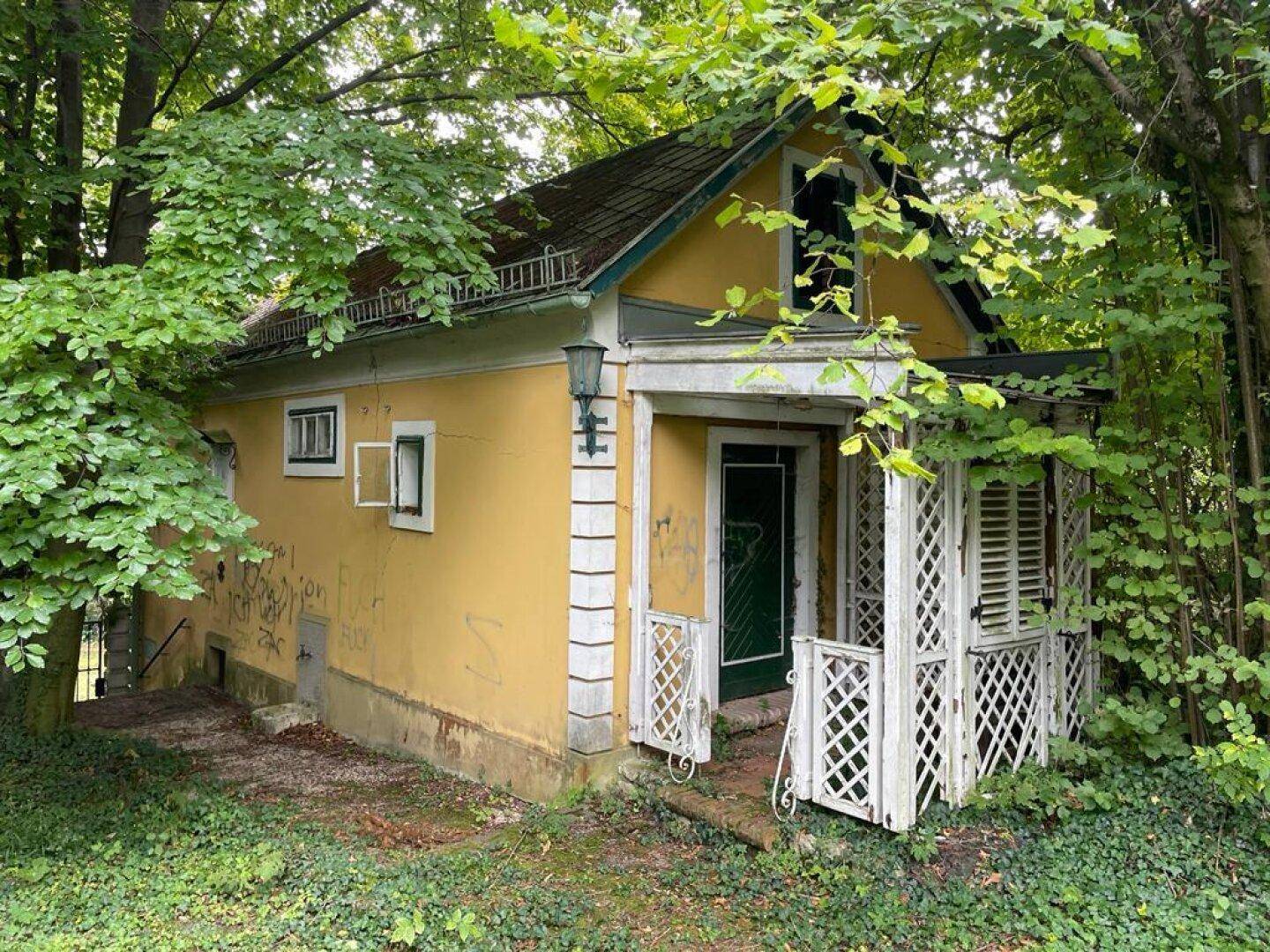 Kleines, älteres Haus mit gelber Fassade und Veranda, umgeben von viel Grün.
