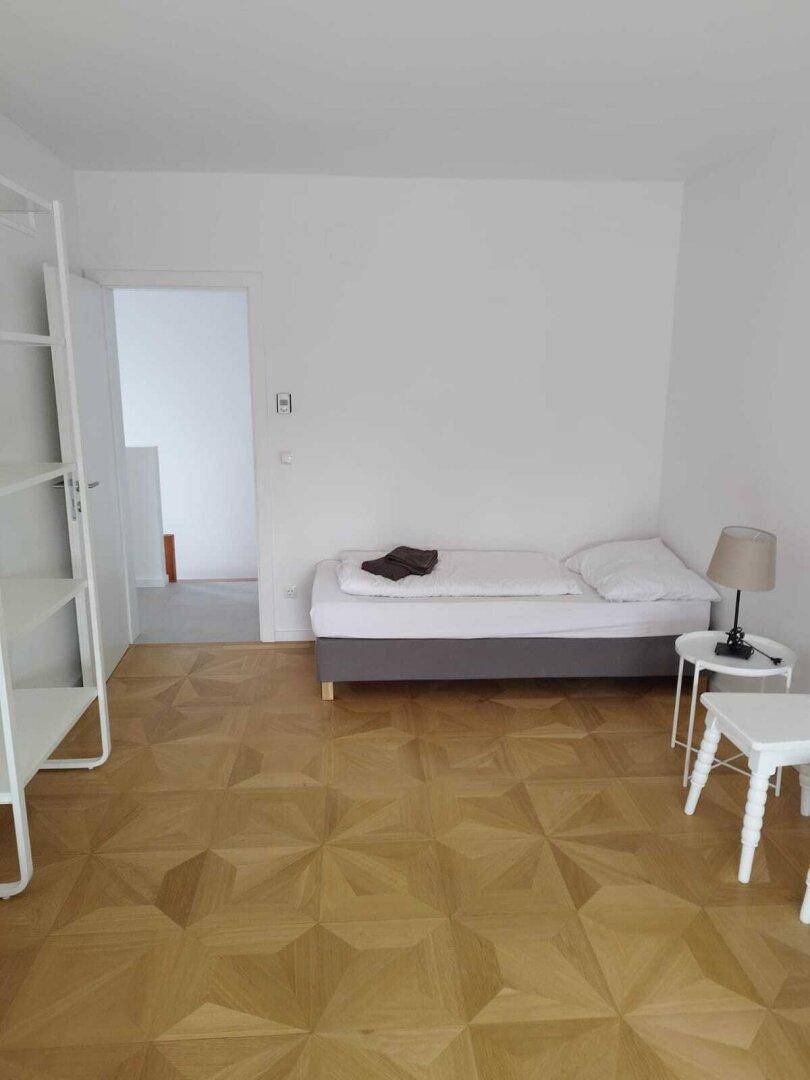 Einfaches Schlafzimmer mit einem Einzelbett, Parkettboden und einem Regal.