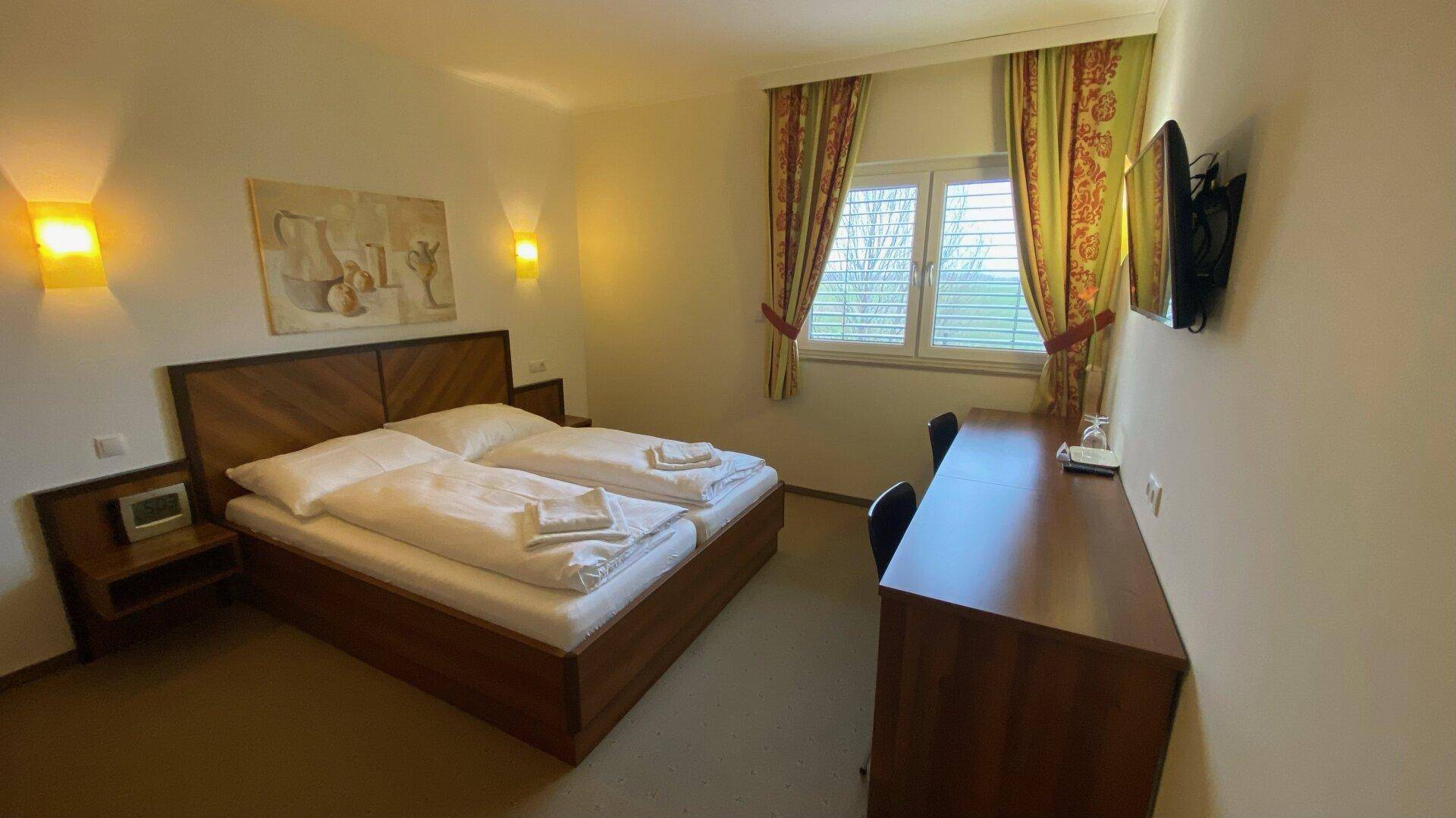 Helles Schlafzimmer mit Doppelbett, Schreibtisch und Blick ins Grüne durch das Fenster.