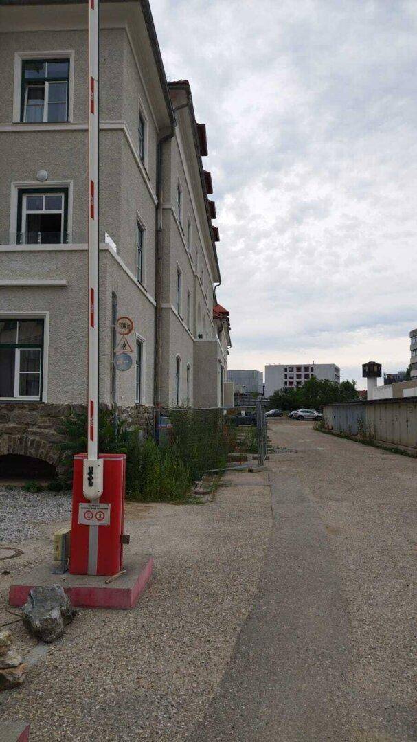 Seitenansicht eines Gebäudes mit Zufahrtsstraße und einer Schranke zur Zugangskontrolle.