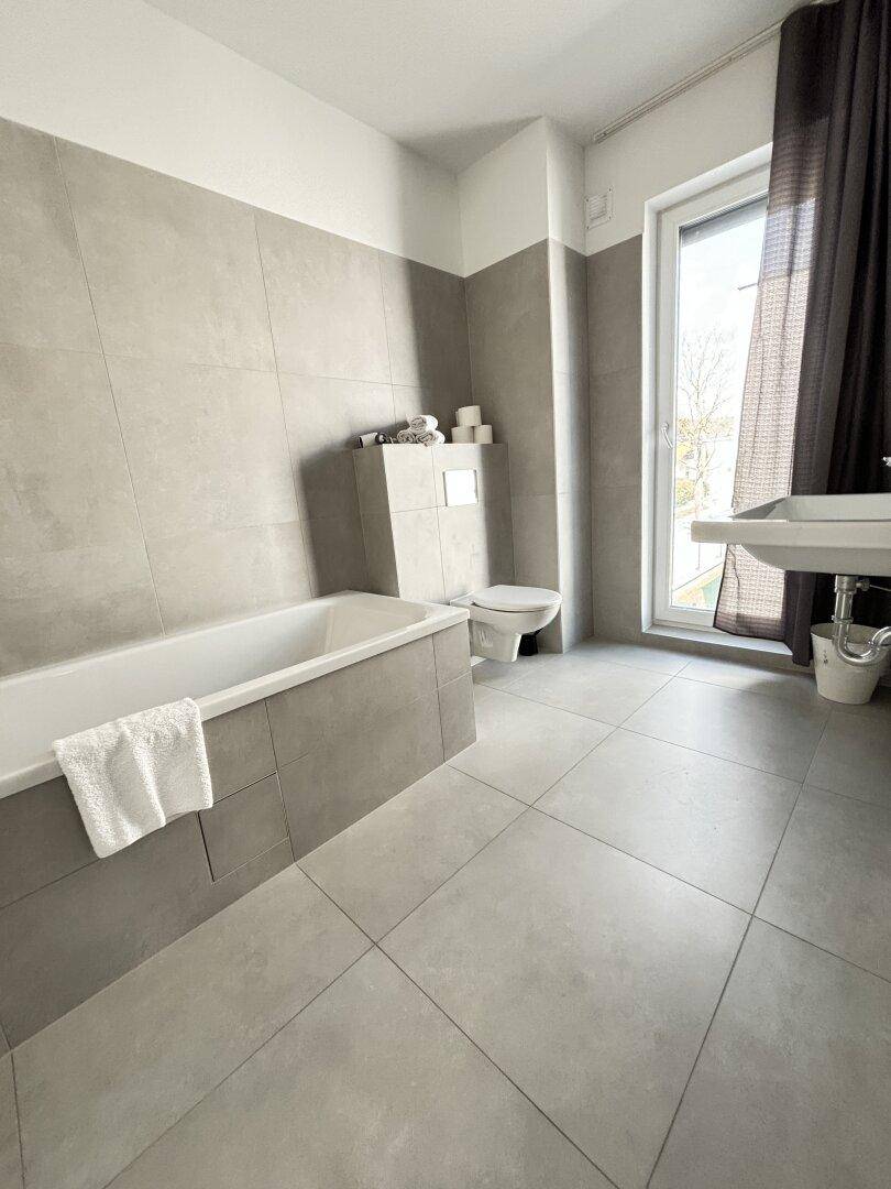 Modernes Badezimmer mit grauen Fliesen, Badewanne, Toilette und Fenster für natürliches Licht.