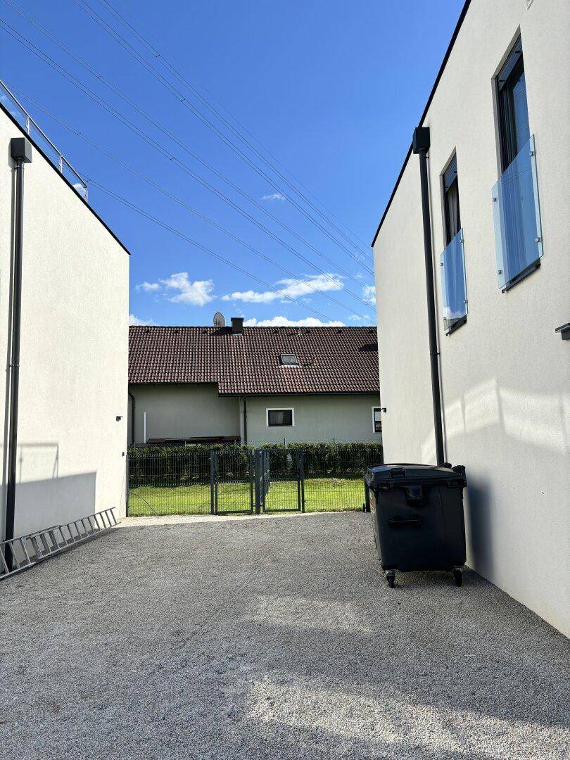 Zwischenraum zweier moderner Gebäude mit Kiesweg und Blick auf ein traditionelles Haus im Hintergrund.
