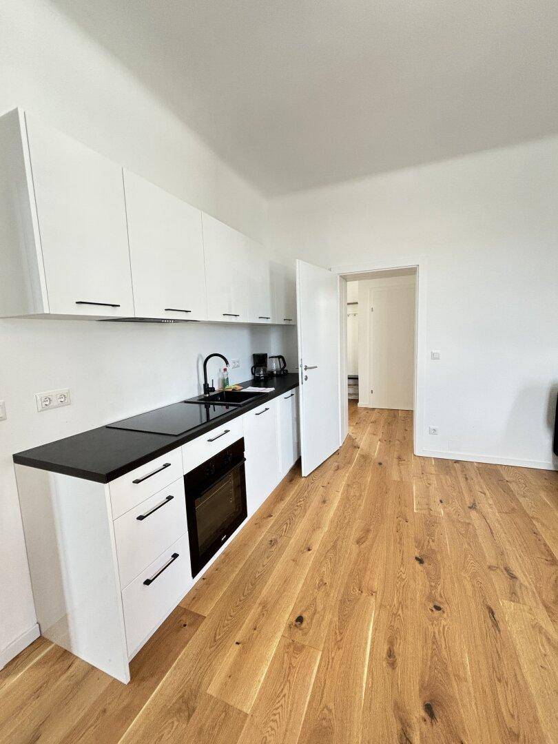 Moderne Küchenzeile mit weißen Schränken, schwarzer Arbeitsplatte und Holzboden in einem hellen Raum.