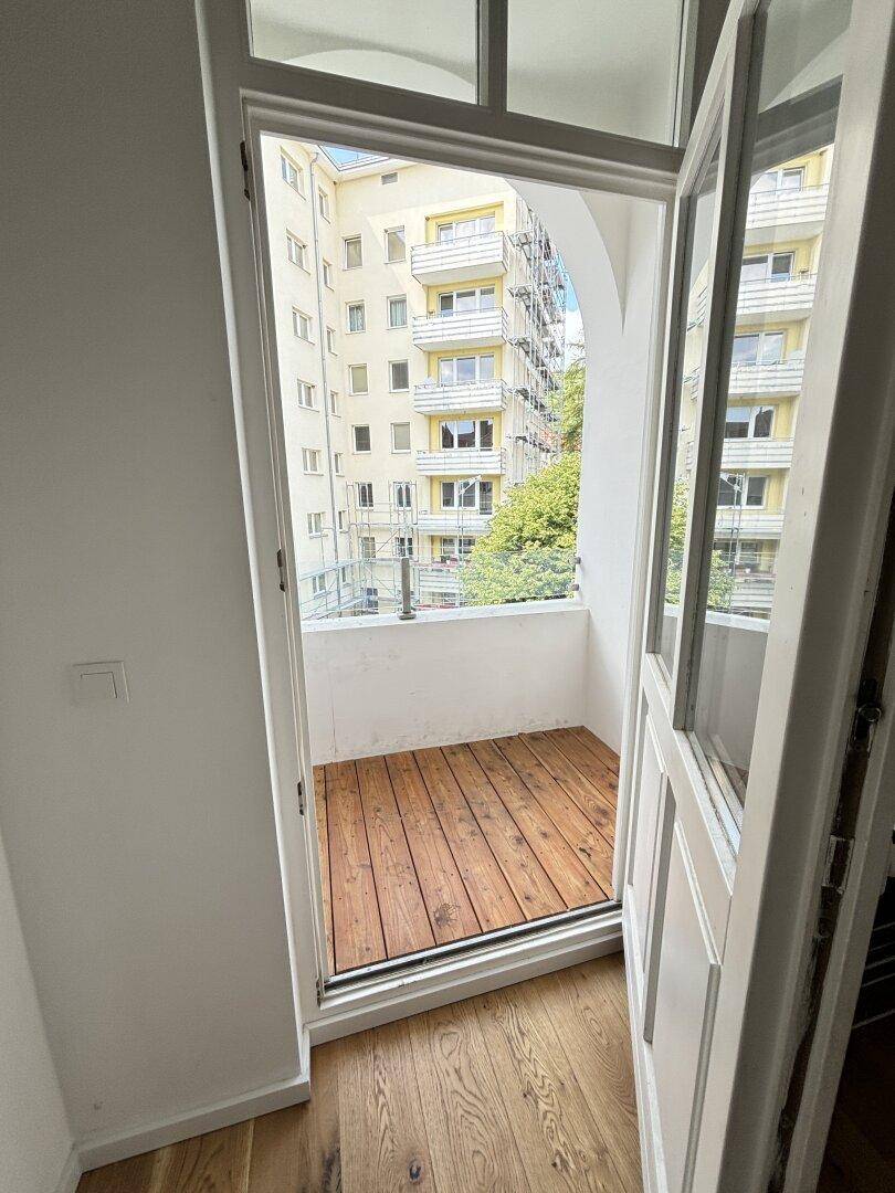 Kleiner Balkon mit Holzboden und Blick auf umliegende Gebäude und Bäume.