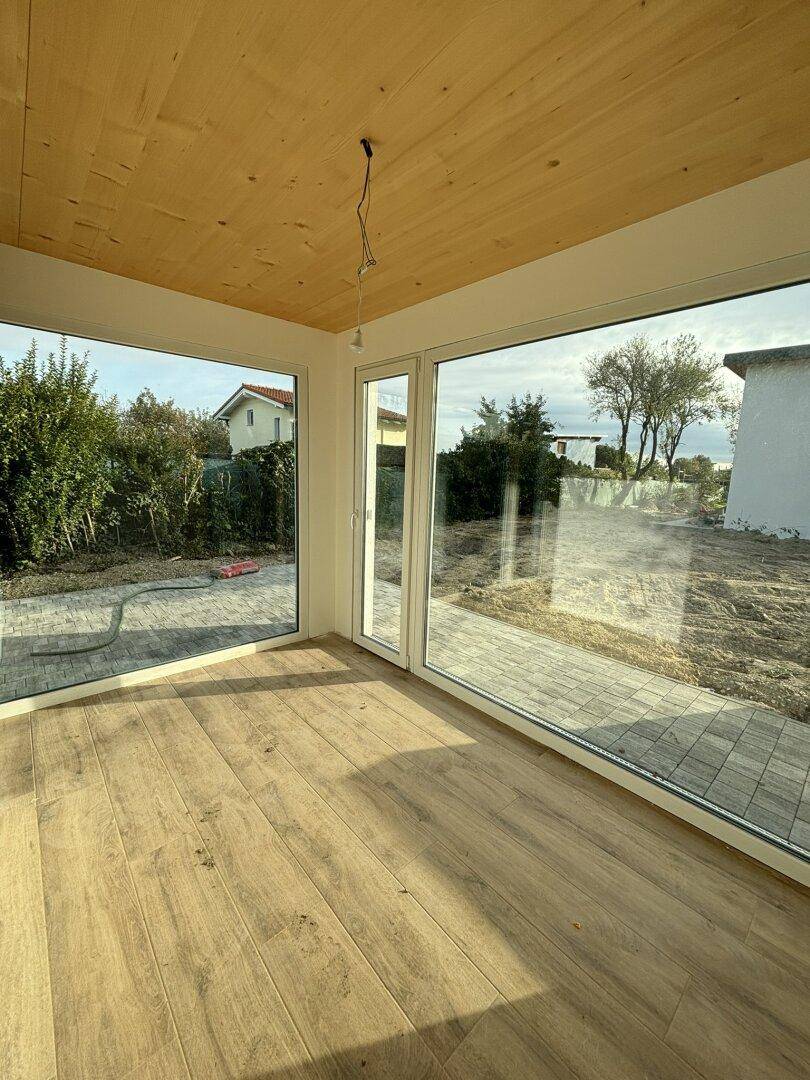 Heller Raum im Rohbau mit Holzboden, Holzdecke und großen Fenstern zum Garten.