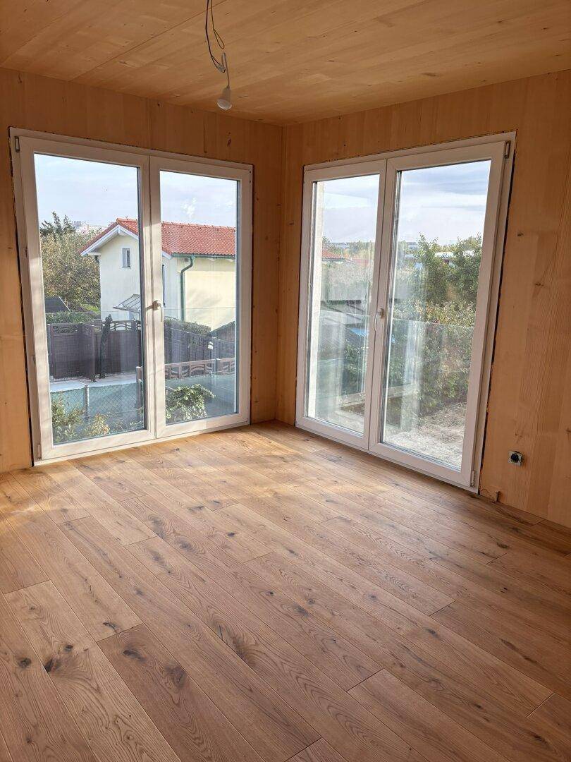 Unfertiger Raum mit Holzboden und Holzdecke, ausgestattet mit großen Fenstern.