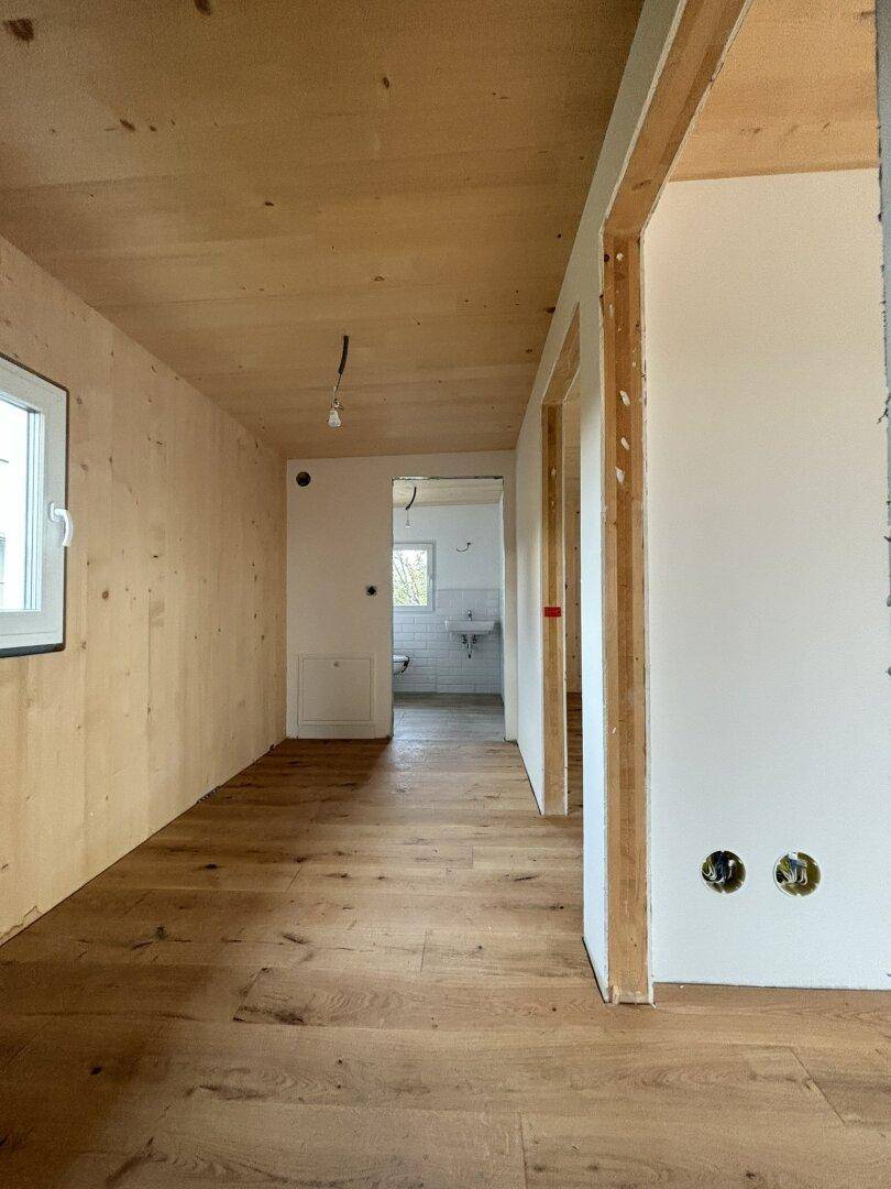 Flur im Rohbau mit Holzwänden, Holzdecke und Holzboden, führt zu einem Badezimmer.