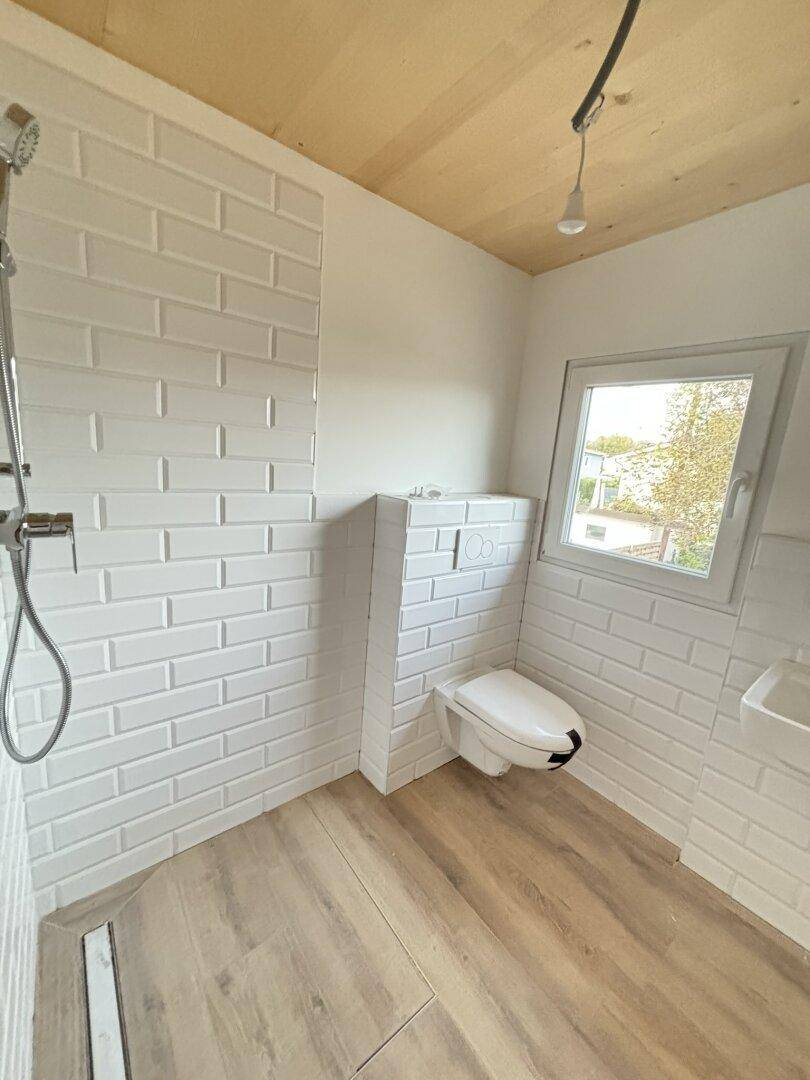 Modernes Badezimmer im Rohbau mit weißen Fliesen, Dusche und wandhängendem WC.