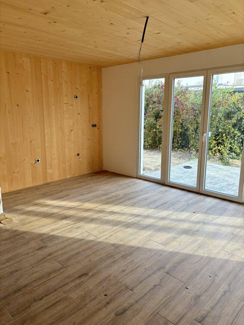 Heller Raum im Rohbau mit Holzwand, Fliesenboden und großen Fenstern zum Außenbereich.