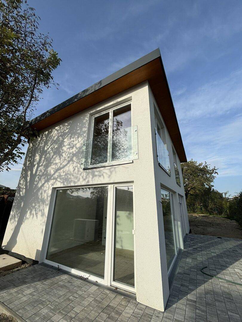 Eckansicht des modernen Hauses mit großen Glasflächen und gepflastertem Bereich im Freien.