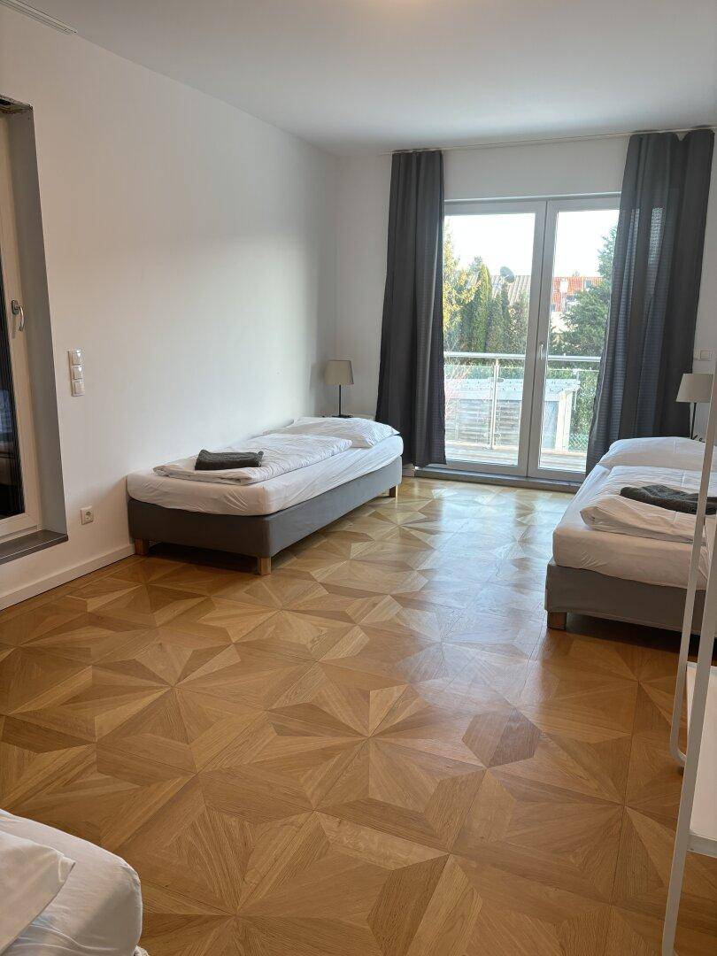 Helles Schlafzimmer mit zwei Betten, Parkettboden und Zugang zu einem Balkon.