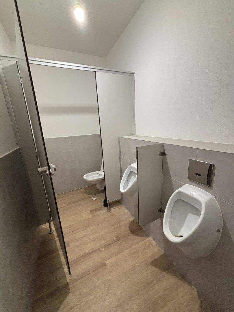 Sauberer und moderner Waschraum mit mehreren Urinalen und einer Toilettenkabine.
