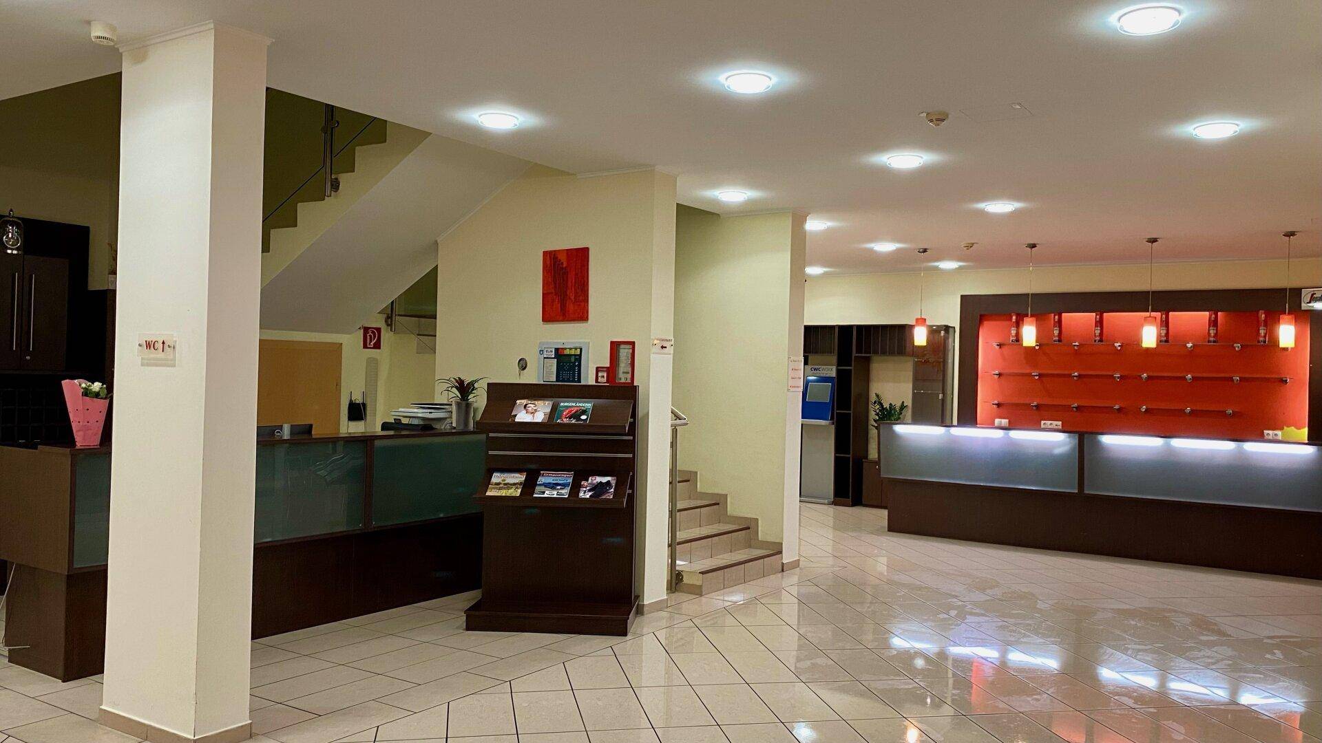 Geräumige Hotellobby mit Rezeption, Informationsständer und einer Treppe, die in die oberen Etagen führt.