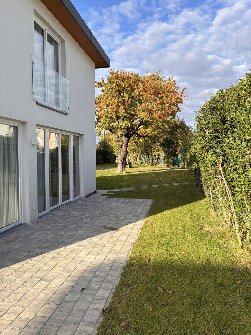 Gepflasterte Terrasse neben dem Haus, angrenzend an den Rasen und eine Hecke mit Herbstbäumen.