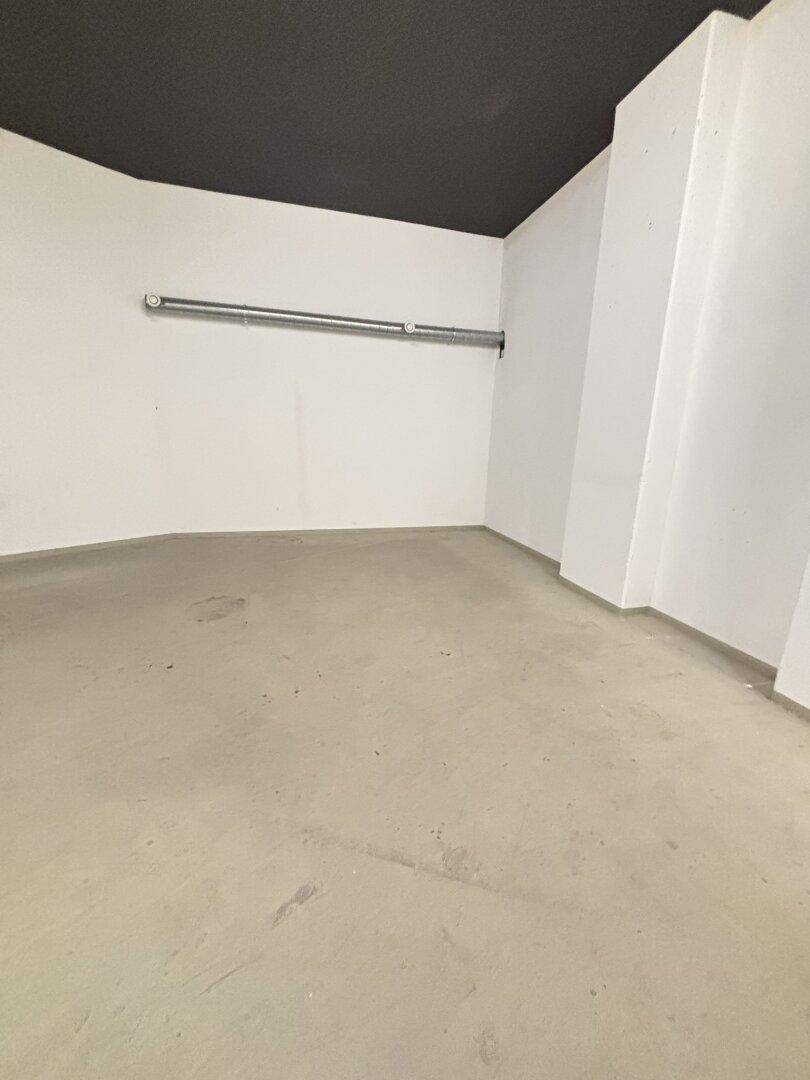 Einfacher Raum mit Betonboden, weißen Wänden und einer sichtbaren Rohrleitung an der Wand.