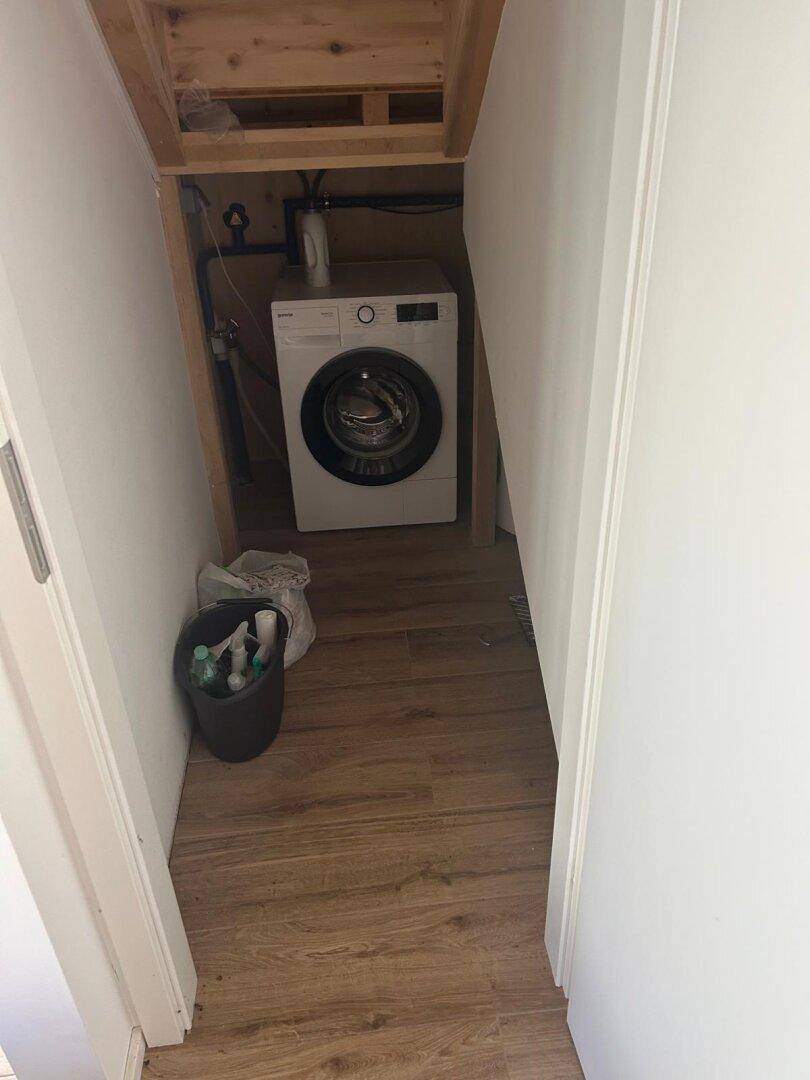 Funktionaler Waschraum unter einer Treppe mit Waschmaschine, Holzverkleidung und Laminatboden.