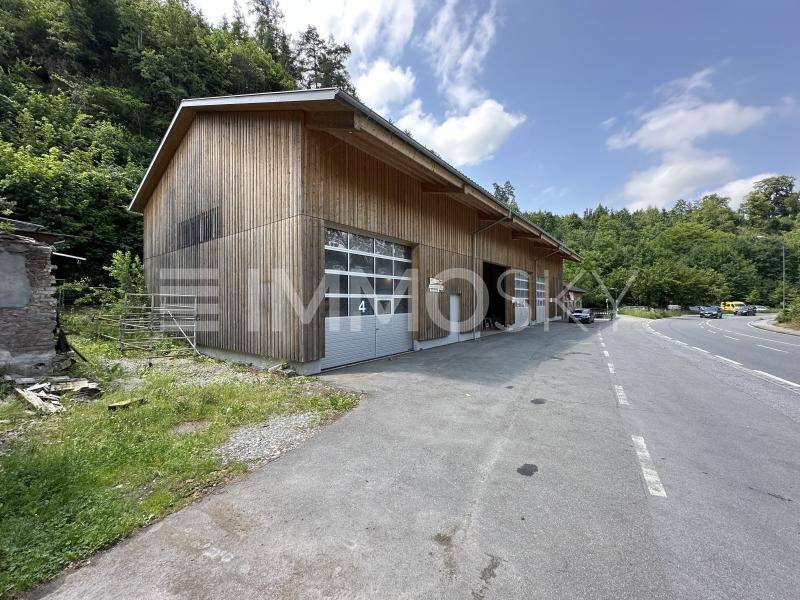 Außenansicht eines modernen Holzgebäudes mit Rolltoren an einer Straße, umgeben von grünen Hügeln.